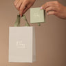 Hands holding a green box and white bag with 'Lily charmed' branding against a brown background