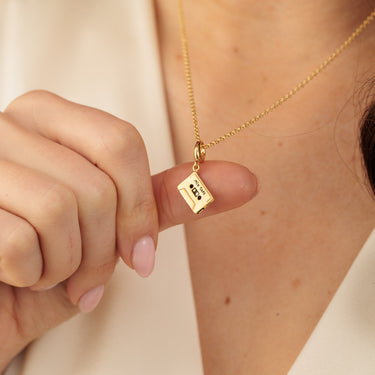 Gold cassette tape necklace held by a hand against a neutral background